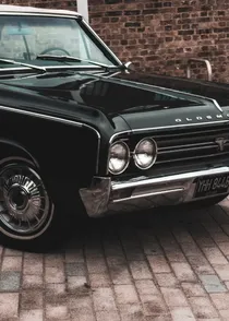 Front of a black vintage Oldsmobile Cutlass parked against a brick wall