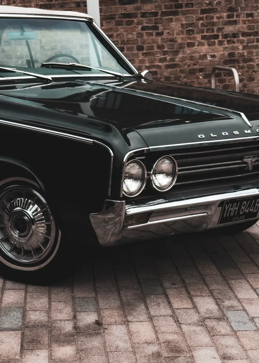 Front of a black vintage Oldsmobile Cutlass parked against a brick wall