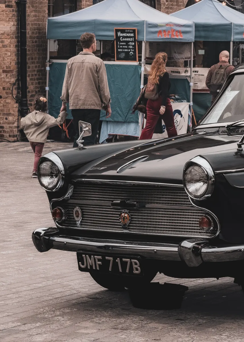 Front of a black vintage car at a street market, with shoppers browsing stalls behind it